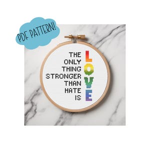 May include: A cross-stitch embroidery in a wooden hoop. The design features the words "THE ONLY THING STRONGER THAN HATE IS LOVE" in black and rainbow colors. A blue cloud-shaped text bubble says "PDF PATTERN!"