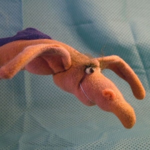 May include: A pink and purple felt puppet shaped like an aardvark with a single black eye and a long snout.
