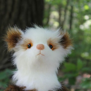 Puede incluir: Un juguete de peluche blanco y marrón con grandes ojos redondos de color naranja. El juguete tiene una cara blanca y esponjosa y pelaje marrón alrededor de las orejas y el cuerpo.