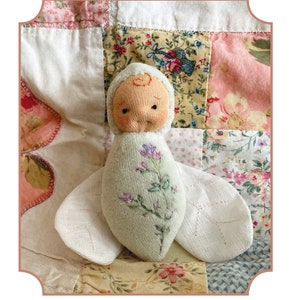 Könnte beinhalten: Eine kleine, handgefertigte Waldorf-Taschenpuppe mit einem hellgrünen Körper und gestickten Blumen. Die Puppe liegt auf einem Patchwork-Deckenstück mit Blumenmustern.