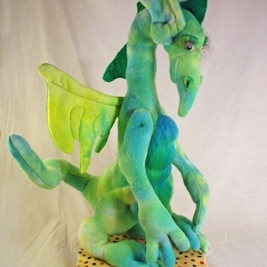 May include: A green and blue plush dragon with a yellow wing, sitting on a patterned fabric. The dragon has a long snout and a single eye.