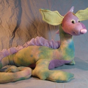 May include: A plush sea dragon toy with a green, purple, and yellow color scheme. The dragon has a long tail, a frilly purple fin, and large yellow ears.