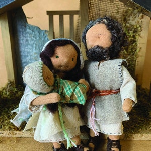 May include: A nativity scene with three handmade dolls. The dolls are dressed in simple brown and white clothing. The mother doll is holding a baby doll wrapped in a green and white blanket. The father doll has a brown beard and is standing next to the mother doll.