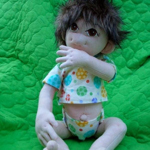May include: A handmade doll with brown hair, wearing a white and green polka dot shirt and matching diaper. The doll is sitting on a green quilted fabric.