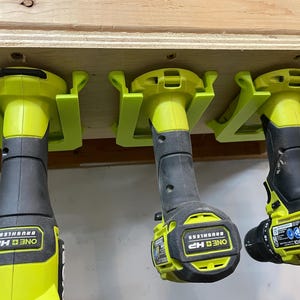 May include: Three green and black cordless drills are mounted on a wall using a 3D printed drill holder. The drills are labeled "ONE+ HP BRUSHLESS".