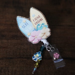 May include: A blue and white glitter badge reel with a surfboard design. The surfboard says "Sun Surf" and "I Love Ocean". The reel has a pink flower charm and a blue flower charm.
