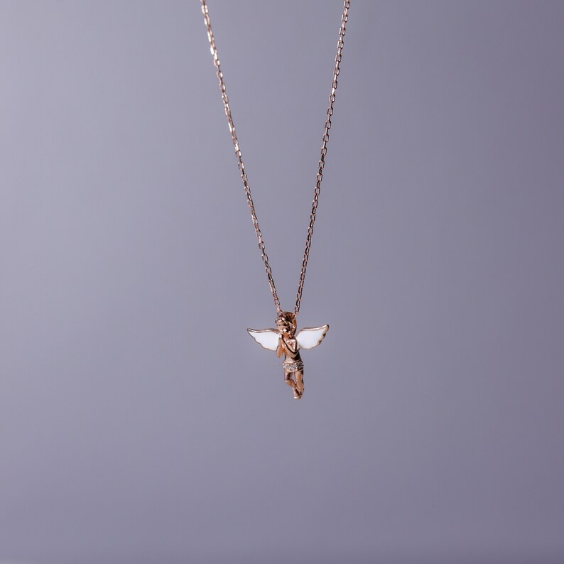 Puede incluir: Un collar de cadena de oro con un peque&ntilde;o colgante de &aacute;ngel. El &aacute;ngel tiene alas blancas y tiene las manos juntas en oraci&oacute;n.