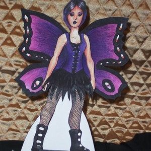 May include: A paper cutout of a fairy with purple butterfly wings. The fairy wears a purple corset top, a black skirt, and black boots. The wings are purple and pink with black outlines. The fairy is standing on a white base.
