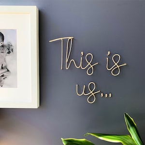 May include: Gold wire wall art with the words "This is us..." on a grey wall.
