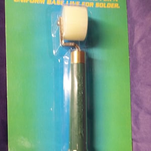 May include: A green and gold hand-held roller with a white plastic roller head. The roller is packaged on a blue and green card with the text "UNIFORM BASE LINE FOR SOLDERING".