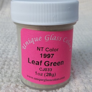 May include: A small glass jar with a white lid and a pink label. The label says "Unique Glass Colors", "NT Color 1997 Leaf Green", "CJ033", "1 oz (28g)", and "www.uniqueglasscolors.com".