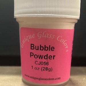 May include: A small, clear plastic jar with a white lid, containing bubble powder. The jar has a pink label with the text "Bubble Powder" and "Unique Glass Colors." The label also indicates the contents are 1 oz (28g).