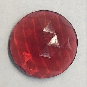 May include: A round, faceted, red gemstone with a reflective surface. The gem has a circular shape and appears to be made of a translucent material. The facets create a play of light and shadow across the surface.