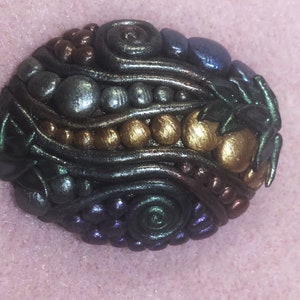 May include: A dark gray, iridescent, oval-shaped polymer clay bead with a textured surface. The bead features a design of swirls, beads, and leaves in shades of gold, silver, green, and blue.
