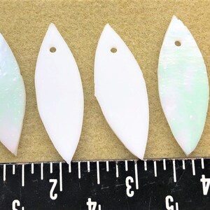 May include: Six leaf-shaped pendants with a hole at the top. The pendants are white, with some featuring pink and green iridescent accents. A ruler is in the foreground.