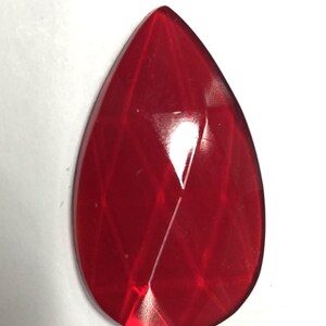 May include: A faceted, teardrop-shaped, red gemstone. The gem has a glossy finish and a series of intersecting lines that create a diamond pattern across its surface. The gem is set against a white background.