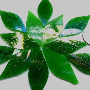 May include: A collection of ten leaf-shaped green glass mosaic tiles arranged in a circular pattern. The tiles feature a glossy finish with subtle white and gold-toned flecks. A small circular metal coin is placed next to the tiles for scale.