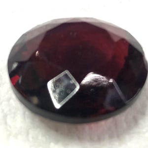 May include: A deep red oval gemstone with a faceted cut. The gemstone has a high polish and is set in a clear, faceted setting.