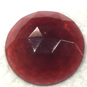 May include: A deep red, faceted, round glass cabochon. The cabochon has a smooth, polished surface and a slightly domed shape.