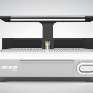May include: A silver and black electric grinder with a white surface and a light above. The grinder has a control panel with two buttons and the text "THE GRINDER TECH-LAB" on the front.
