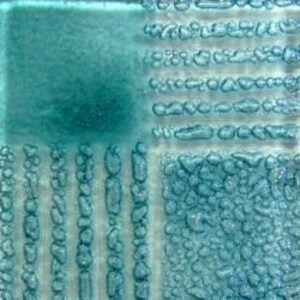 May include: A close-up of a teal blue glass tile with a textured surface. The tile has a smooth, solid teal blue section and a section with raised, bubble-like textures in various sizes and patterns.