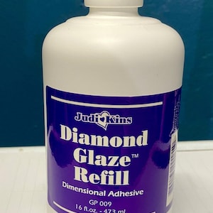 May include: A white plastic bottle with a white cap, featuring a purple label with the text "Judi Kins Diamond Glaze Refill." The label also indicates the product is a dimensional adhesive, with a volume of 16 fl. oz. - 473 mL.