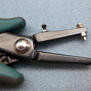 May include: A black and green metal hand punch tool with a silver punch head. The tool is used for making holes in leather, paper, or other materials.