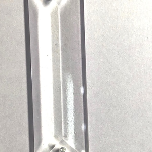 May include: A clear, rectangular glass prism with beveled edges and two small, circular metal attachment points located near the top and bottom ends. The object features a smooth, transparent surface that reflects light across its elongated body.