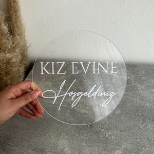 May include: A clear, round acrylic sign with white text that reads "KIZ EVINE Hosgeldiniz" in a mix of block and script fonts. The sign is held by a hand against a gray surface, with dried plants in the background.