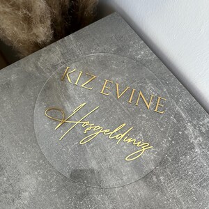 May include: A clear, circular sign with gold lettering. The text reads "KIZ EVINE Hosgeldiniz" in a decorative font. The sign is placed on a gray, textured surface, with dried plants in the background.