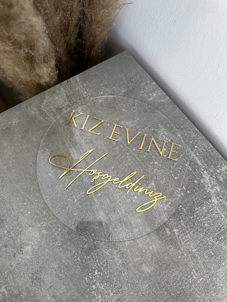 May include: A clear, circular sign with gold lettering. The text reads "KIZ EVINE Hosgeldiniz" in a decorative font. The sign is placed on a gray, textured surface, with dried plants in the background.