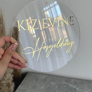 May include: A clear, round sign with gold lettering. The text reads "KIZ EVINE Hosgeldiniz". The sign has a small hole at the top for hanging. The sign is held by a hand.