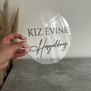 May include: A clear, round acrylic sign with the words "KIZ EVINE Hosgeldiniz" in black script. The sign has a small hole at the top for hanging. The sign is held in front of a gray surface.