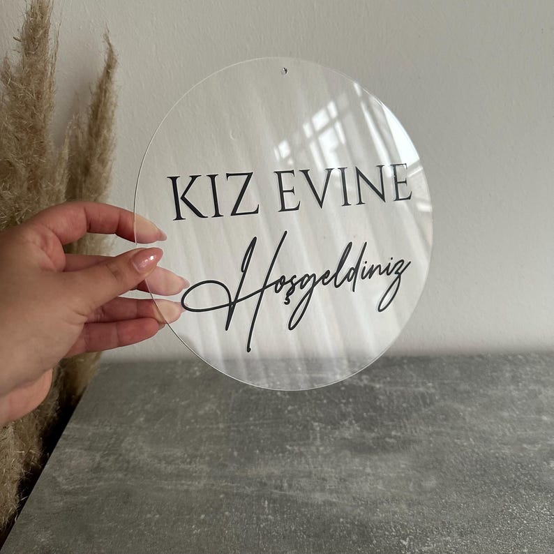May include: A clear, round acrylic sign with the words "KIZ EVINE Hosgeldiniz" in black script. The sign has a small hole at the top for hanging. The sign is held in front of a gray surface.