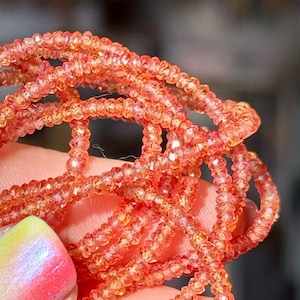 Può includere: Primo piano di fili di perline di pietre preziose arancione-rossastro. Le perline sono piccole, sfaccettate e infilate insieme, creando un effetto scintillante. Le perline sono tenute in una mano con una manicure colorata.