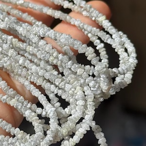 Könnte beinhalten: Eine Strähne aus rohen, weißen Diamantperlen. Die Perlen sind klein und unregelmäßig geformt und auf einem durchsichtigen Faden aufgereiht. Die Perlen sind in einem lockeren, fließenden Muster angeordnet.