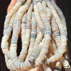 May include: Strands of small, flat, round beads in shades of cream, gray, and pale blue. The beads are strung together, creating a textured and layered effect. The beads are held in a hand.