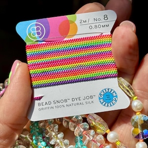 May include: A spool of Bead Snob Dye Job Griffin 100% natural silk cord, size No. 8, 0.80mm, 2 meters long. The cord features a vibrant rainbow-colored gradient pattern. The white cardboard packaging displays the brand logo and product details.