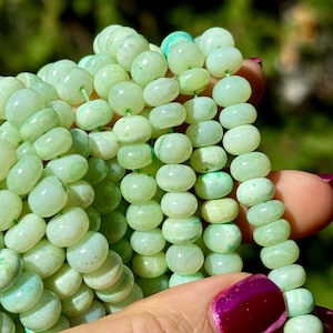 May include: A strand of light green gemstone beads, each bead is round and slightly flattened, with a smooth, polished surface.