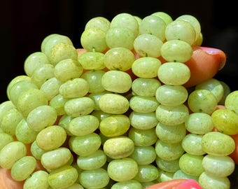 May include: A close-up of a strand of light green, oval-shaped beads. The beads are held in a hand with a painted nail. The beads have a smooth, polished appearance and vary slightly in color, with some appearing more yellow.