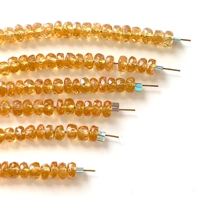 May include: Strands of faceted, golden-yellow gemstone beads. Each bead is strung on a wire, with small, iridescent spacers. The beads are arranged in a diagonal pattern against a white background. Ideal for jewelry making.