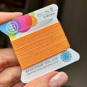 May include: A spool of bright orange Griffin Bead Snob Dye Job 100% natural silk cord. The white cardboard card features the text 2M / No. 8 0.80MM, the Griffin logo, and the product name. The silk cord is wound horizontally across the card.