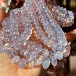 Puede incluir: Primer plano de varias hebras de cuentas de piedras preciosas azul claro y transparentes. Las cuentas de forma ovalada están ensartadas con un hilo fino de color púrpura. Las cuentas translúcidas reflejan la luz.