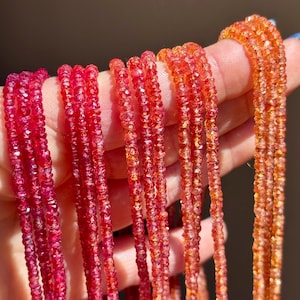 May include: Close-up of several strands of faceted gemstone beads in shades of red and orange. The beads are held up against a blurred background, showcasing their vibrant colours and sparkling texture. The gemstones appear to be small and uniform in size.