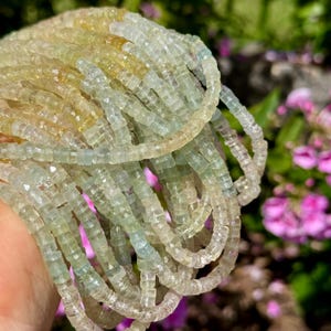 Pu&ograve; includere: Primo piano di pi&ugrave; fili di piccole pietre preziose quadrate in tonalit&agrave; giallo pallido, verde e trasparente. Le perline sono infilate, forse per collane o braccialetti. Lo sfondo &egrave; sfocato con fiori rosa.