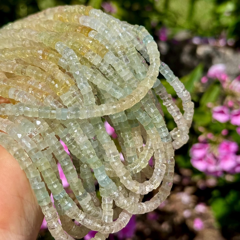 Pu&ograve; includere: Primo piano di pi&ugrave; fili di piccole pietre preziose quadrate in tonalit&agrave; giallo pallido, verde e trasparente. Le perline sono infilate, forse per collane o braccialetti. Lo sfondo &egrave; sfocato con fiori rosa.