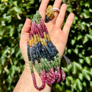 Puede incluir: Una hebra de cuentas de piedras preciosas multicolores, incluyendo amarillo, verde, azul y rosa, enhebradas en un cordón blanco. Las cuentas son facetadas y tienen forma cuadrada.