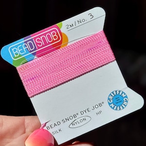 May include: A spool of bright pink nylon thread labeled "BEAD SNOB DYE JOB&reg;". The label also includes the text "SILK" and "NYLON". The thread is 2 meters in length and is marked as No. 3. The label has a colorful design.
