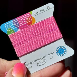 May include: A spool of bright pink nylon thread labeled "BEAD SNOB DYE JOB" and "SILK". The label also includes the text "2M/No. 8" and the Griffin logo. The thread is tightly wound and appears ready for use in jewelry making or crafts.