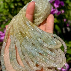 Pu&ograve; includere: Fili di perline di pietre preziose verde chiaro e giallo. Le perline sono piccole, tagliate a forma quadrata e infilate insieme. Le perline sono tenute in una mano, con uno sfondo sfocato di fogliame verde e viola.